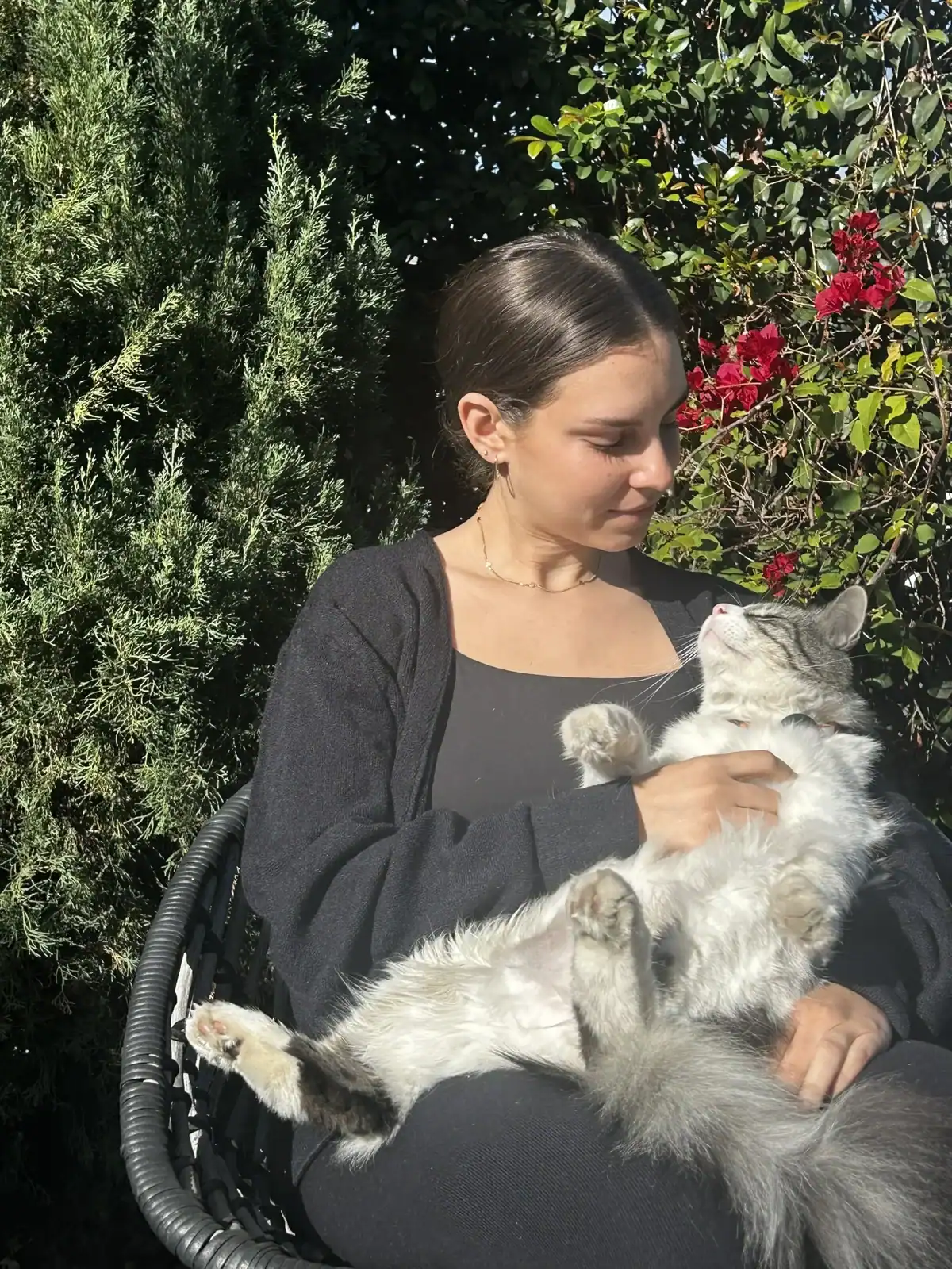 Woman sitting outdoors in a garden chair, gently holding a fluffy gray cat surrounded by green foliage and red flowers.