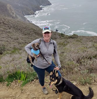 Woman hiking on a coastal trail with two dogs, ocean cliffs visible in the background.
