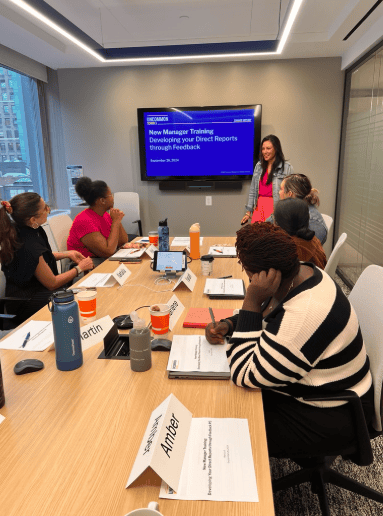 Facilitator leading a small group training session in a conference room with presentation slides on a screen.
