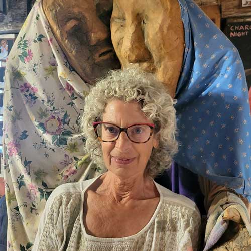 Older woman with curly gray hair and glasses standing in front of large sculptural figures draped in patterned fabric.