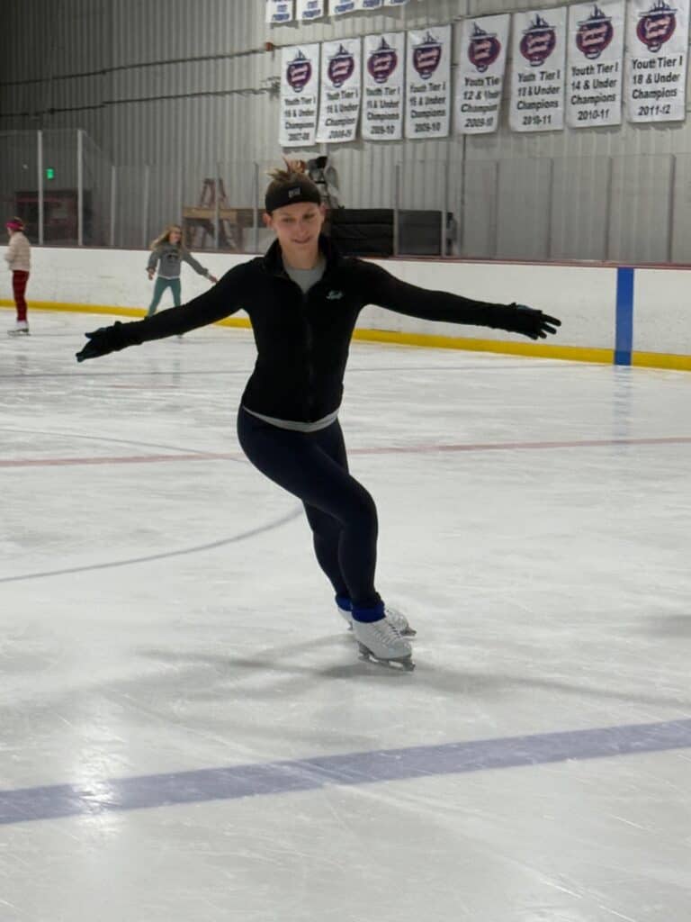 Figure skater in a black jacket, gloves, and leggings skates on an indoor rink with arms out, while other skaters practice in the background.