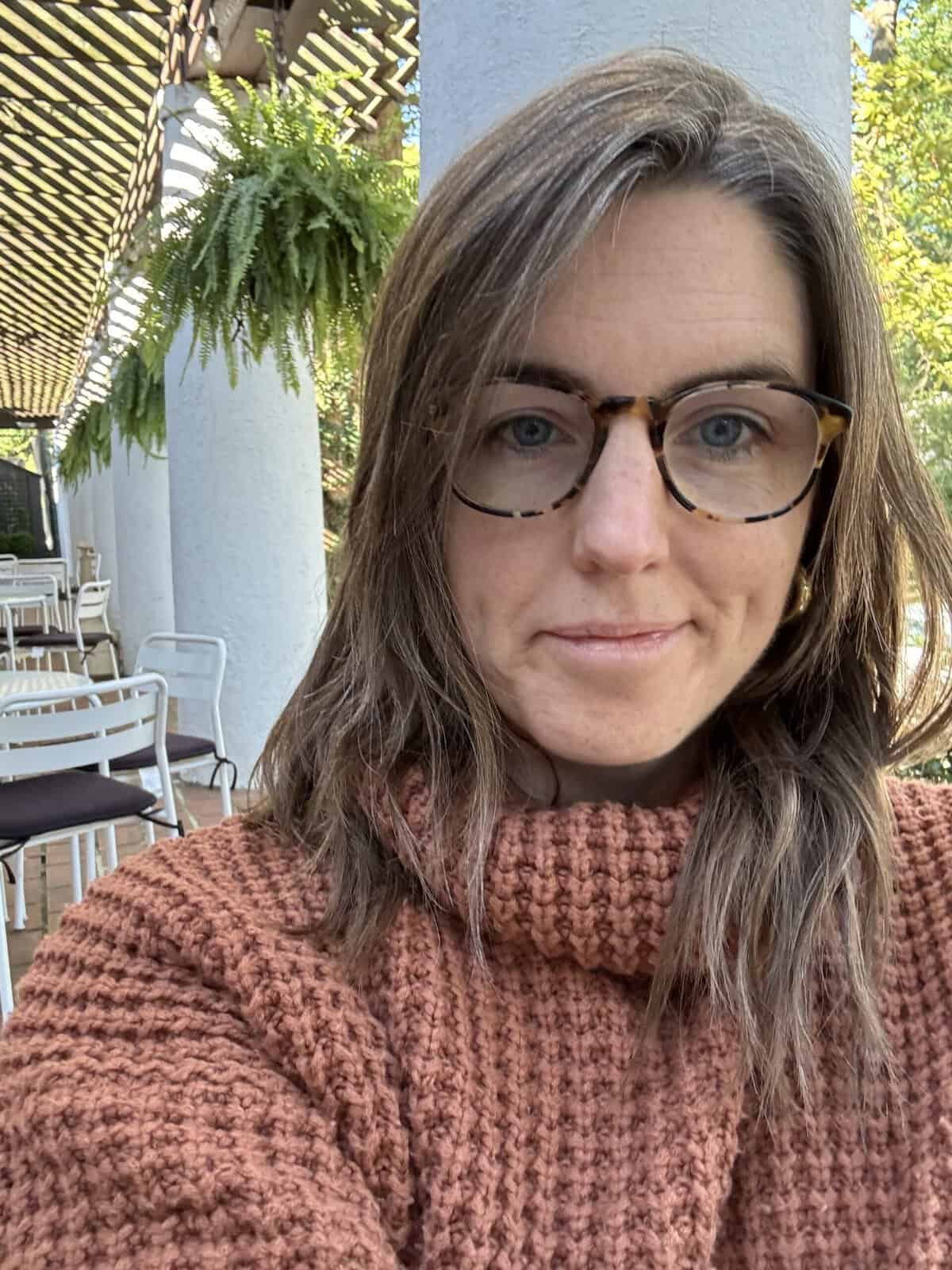 Close-up selfie of a woman wearing glasses and a textured brown sweater, standing outdoors near white columns and hanging plants.