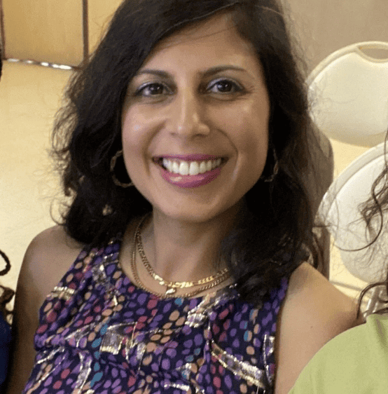 A smiling woman with shoulder-length dark hair looks at the camera. She is wearing a sleeveless purple patterned top, gold hoop earrings, and layered gold necklaces. The background appears to be an indoor event space with neutral-colored walls and stacked chairs.