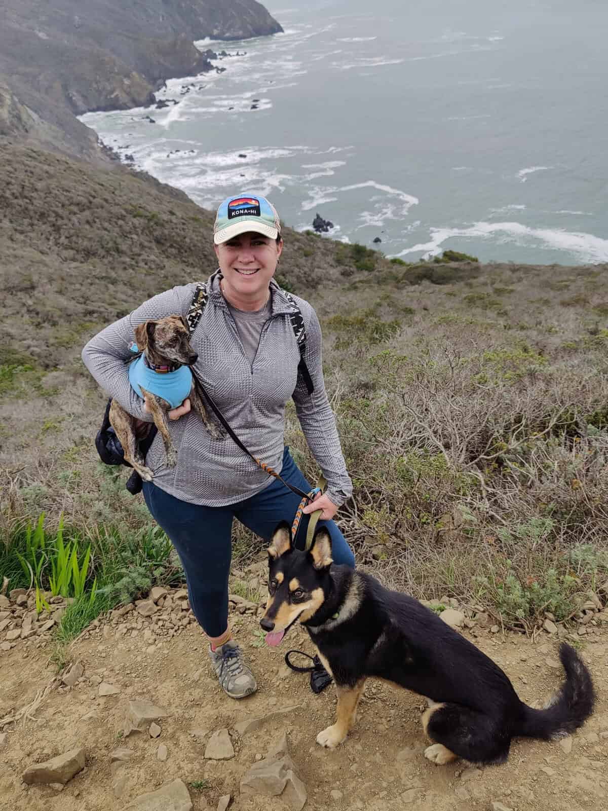 A woman in hiking gear smiles while standing on a dirt trail overlooking a rocky coastline. She is holding a small, brindle dog wearing a light blue sweater under one arm, while her other hand holds the leash of a larger black and tan dog sitting beside her. The background features steep, shrub-covered cliffs meeting a churning gray ocean