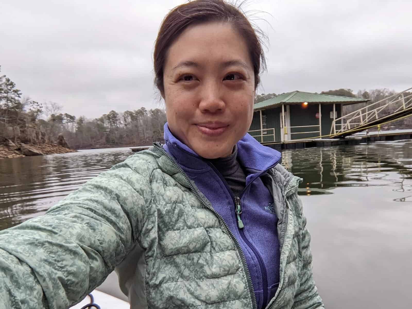Person taking a selfie while sitting on a paddleboard or kayak on a calm lake, wearing a green quilted jacket over a purple layer. A small dock with a covered boathouse and a sloped gangway is visible behind them under an overcast sky.