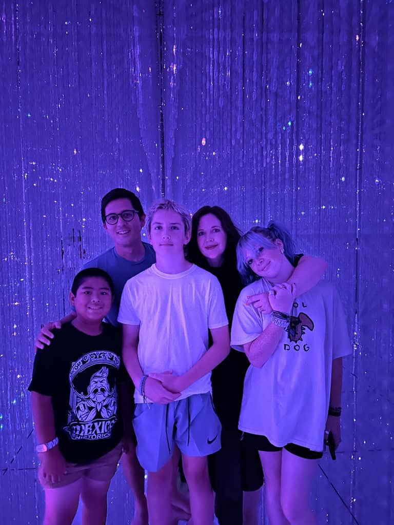 A group of five people posing together in front of a purple-lit, sparkly backdrop.
