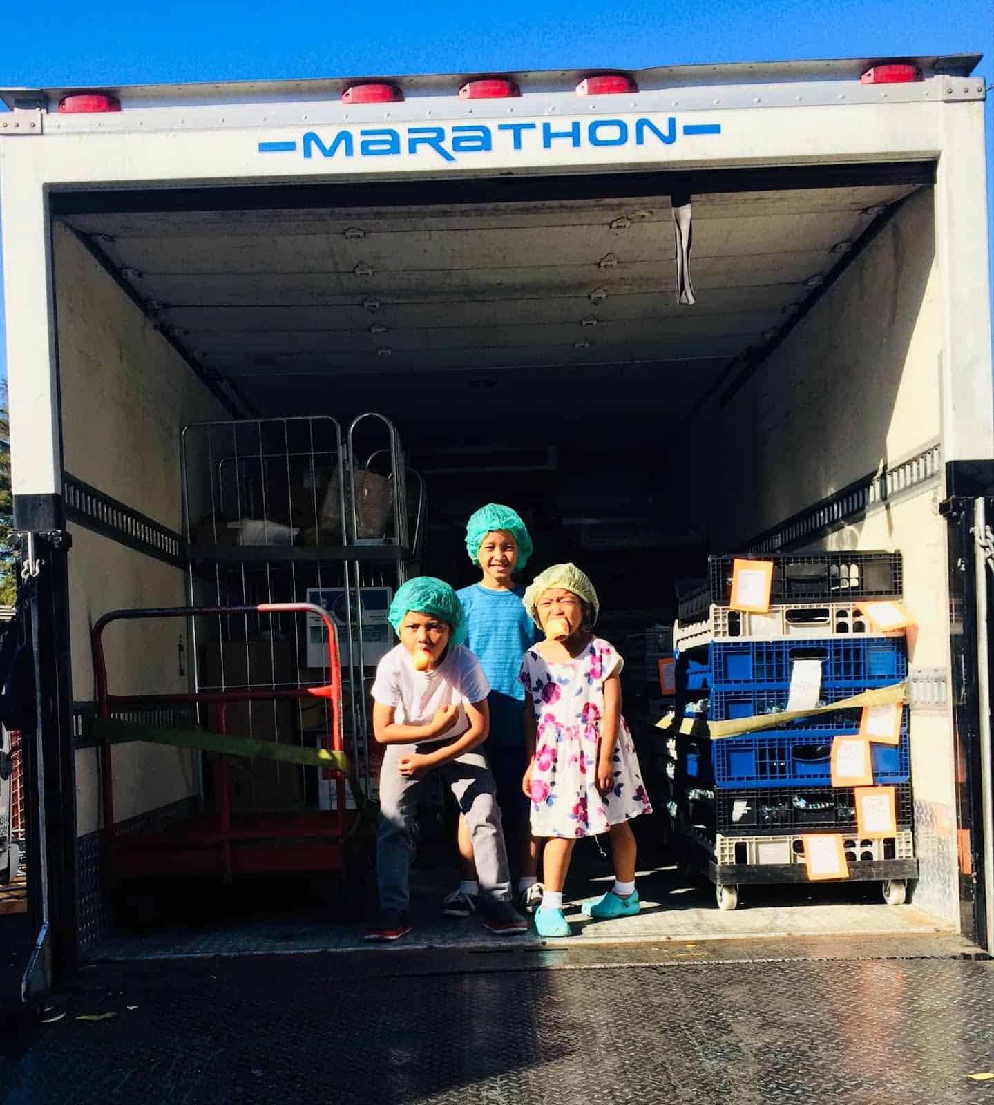 Three children wearing hairnets pose together at the open rear of a Marathon delivery truck.