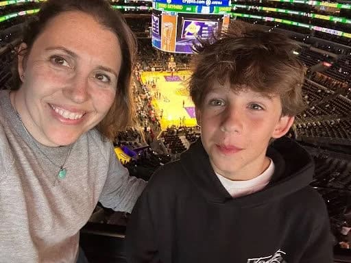 A woman and a preteen boy smile together in a selfie taken from the stands at a basketball arena.
