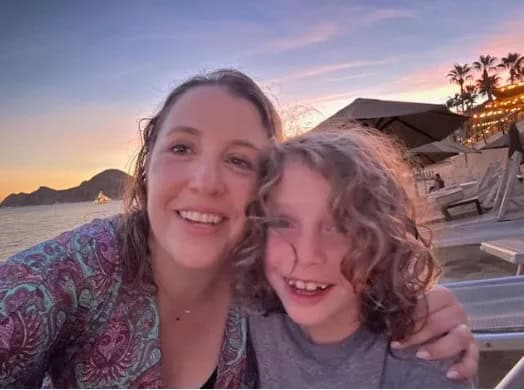 A woman and a young child smile together in a beachside selfie at sunset.