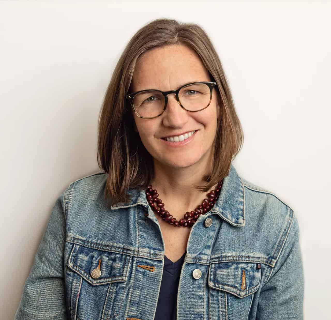 Smiling woman with shoulder-length brown hair wearing glasses, a denim jacket, and a beaded necklace, standing against a plain light background.