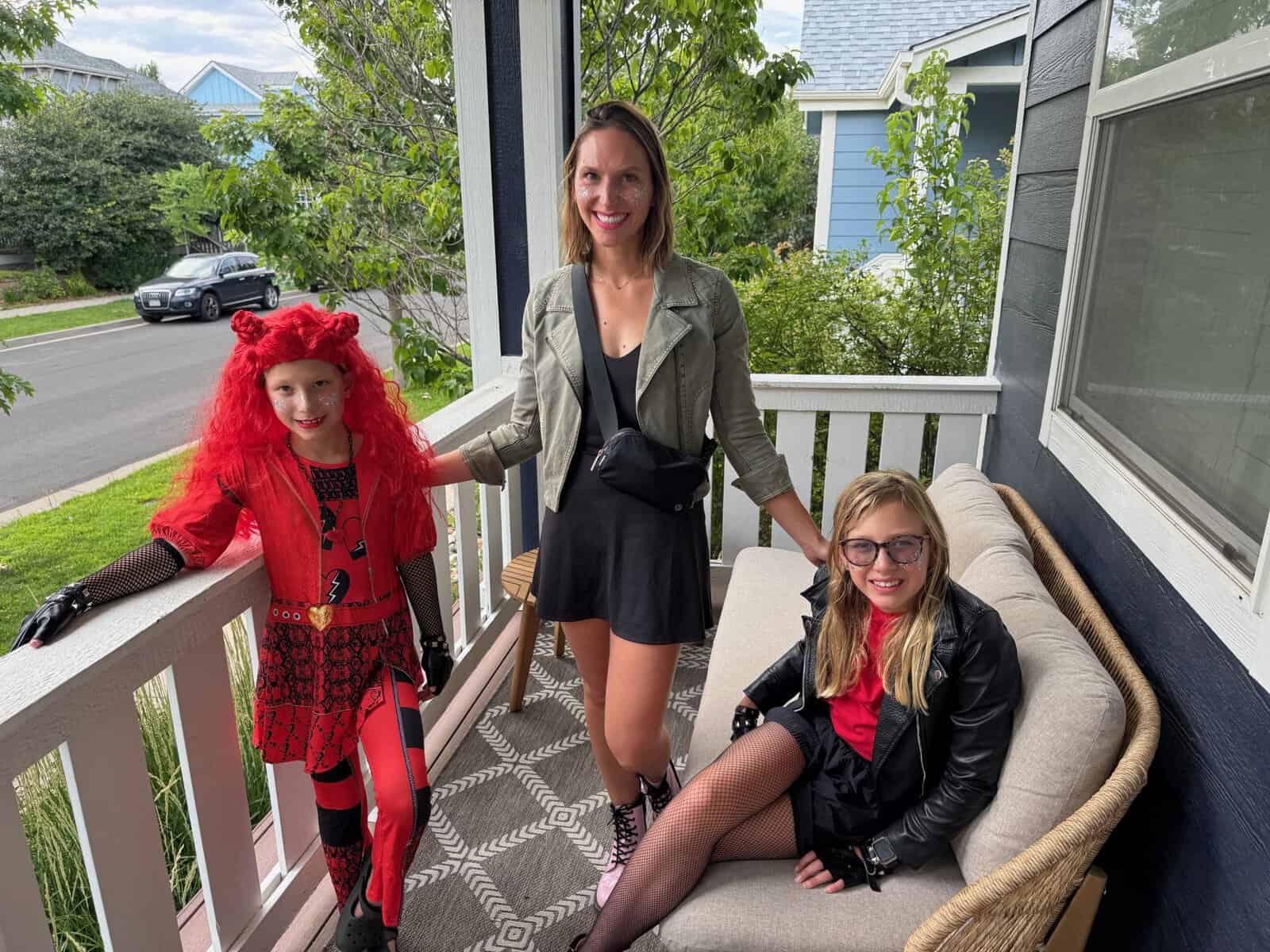 Three people pose together on a covered front porch in a residential neighborhood. An adult woman stands in the center, smiling, wearing a black dress, green jacket, and a crossbody bag. To her left, a child in a bright red costume with long red hair, patterned outfit, and black gloves stands by the railing. To her right, another child sits on a cushioned outdoor bench wearing a black jacket, red top, fishnet tights, and glasses. Trees, neighboring houses, and a parked car are visible in the background.