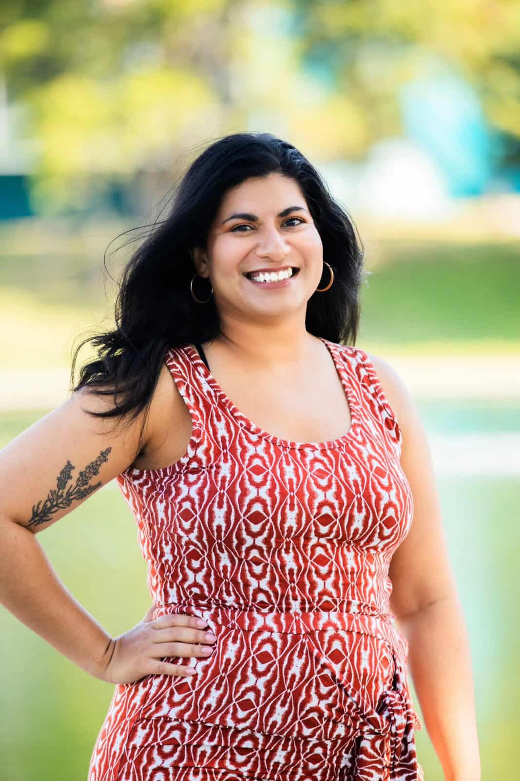 A woman with long, dark wavy hair and gold hoop earrings smiling outdoors. She is wearing a sleeveless red and white patterned dress and has a floral tattoo on her right upper arm. The background is a soft-focus green landscape.