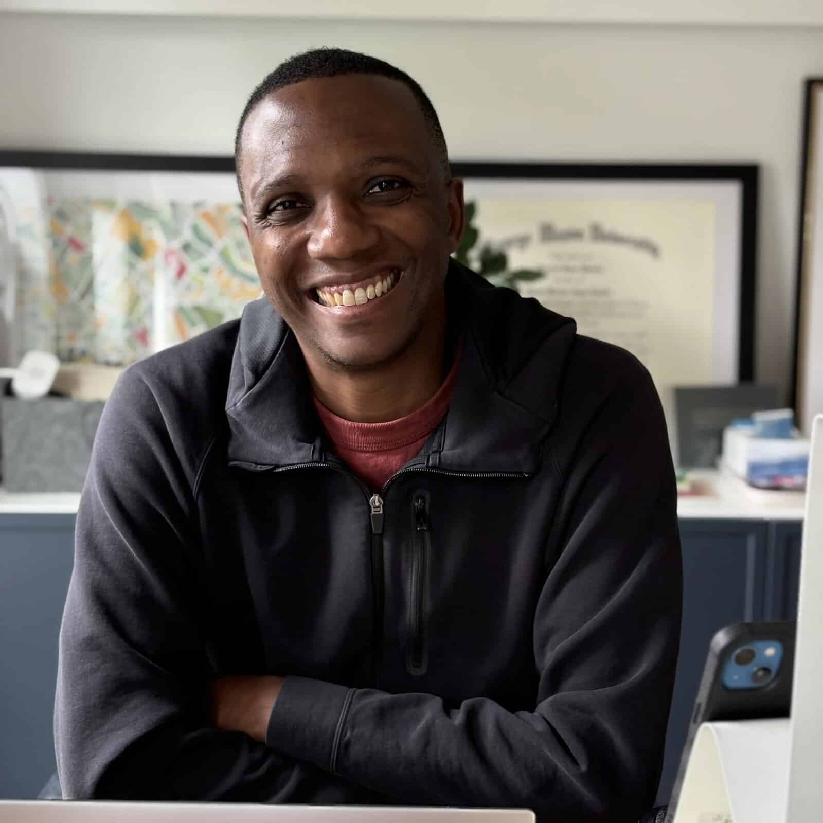 A Black man with a warm, wide smile sits at a desk with his arms crossed, wearing a black zip-up hoodie over a red shirt. He's in a home office setting with framed artwork and a diploma visible in the background.