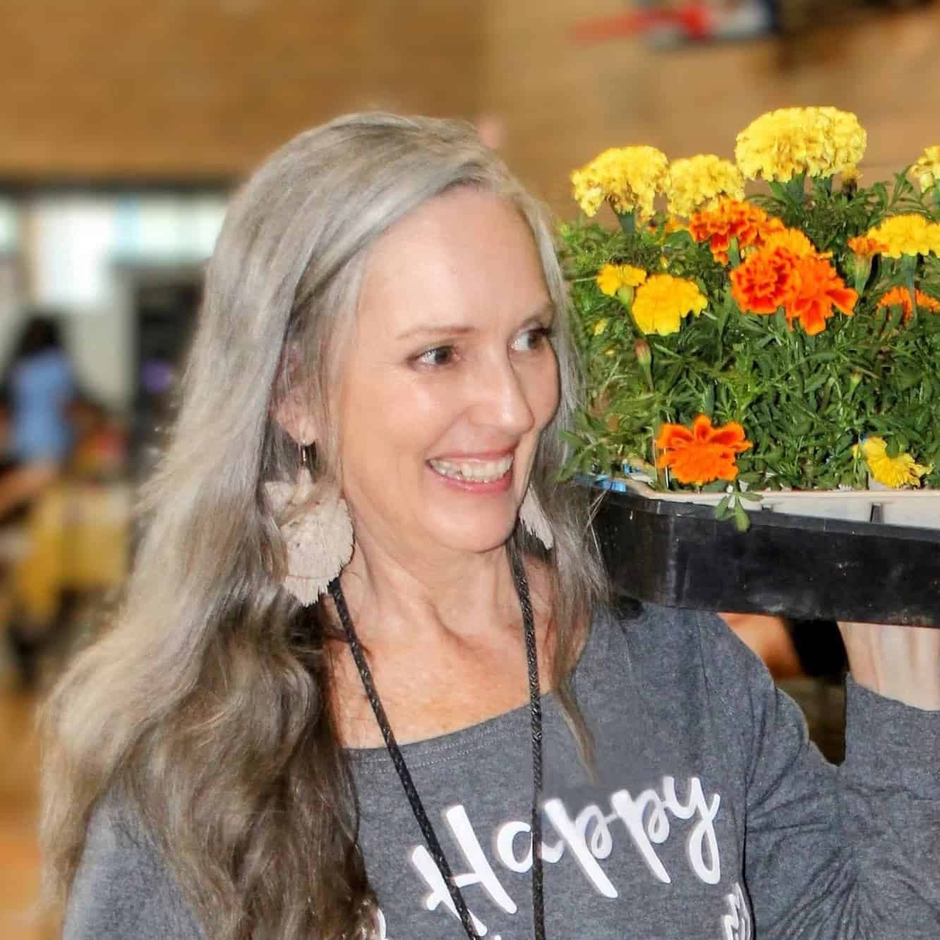 A woman with long, wavy gray hair smiles brightly while carrying a black tray of vibrant yellow and orange flowers on her shoulder. She is wearing a gray long-sleeved shirt with the word "Happy" visible, large tasseled earrings, and a black lanyard.