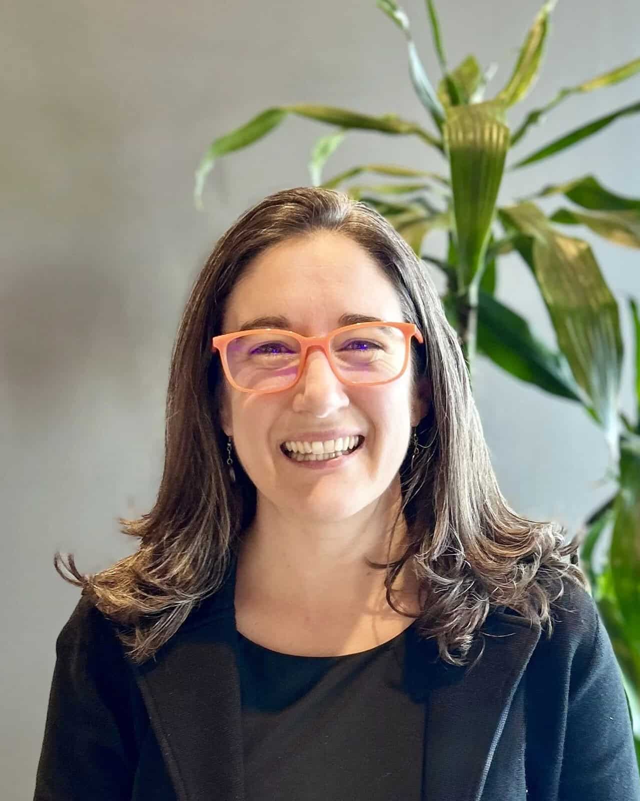 A smiling woman with shoulder-length wavy brown hair and orange-rimmed glasses, wearing a black blazer and standing in front of a green plant and a gray wall.