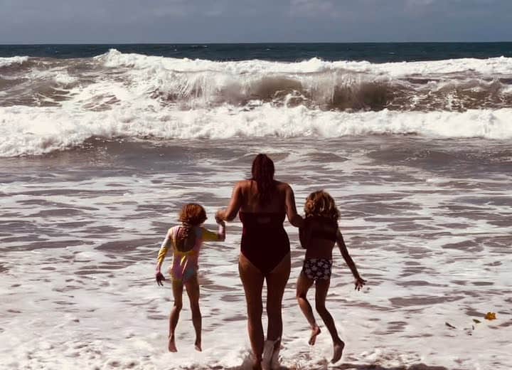 A woman holding hands with two young girls, seen from behind, facing crashing ocean waves on a sandy beach