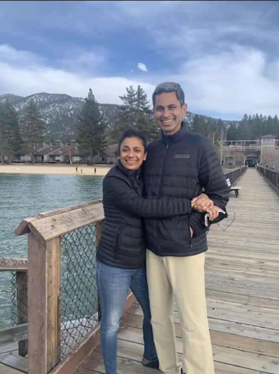 Smiling couple standing on a wooden pier by a lake, with mountains and cabins in the background.