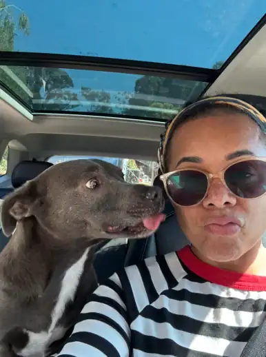 Woman taking a selfie in a car while her dog leans in and licks her face.