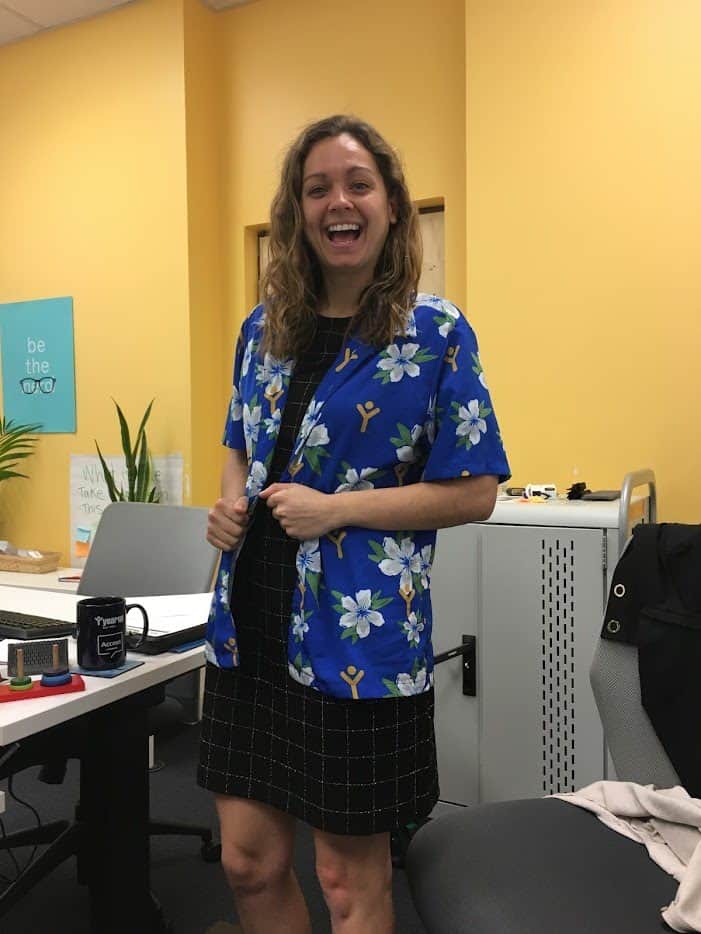 A woman stands in an office, smiling brightly while wearing a colorful blue floral shirt over a dress. The space feels casual and friendly, with a relaxed, upbeat vibe.
