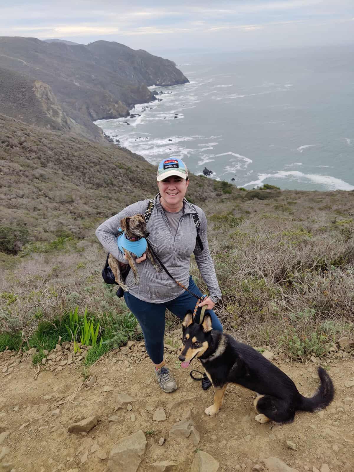 Woman hiking on a coastal trail with two dogs, overlooking rugged cliffs and the ocean.