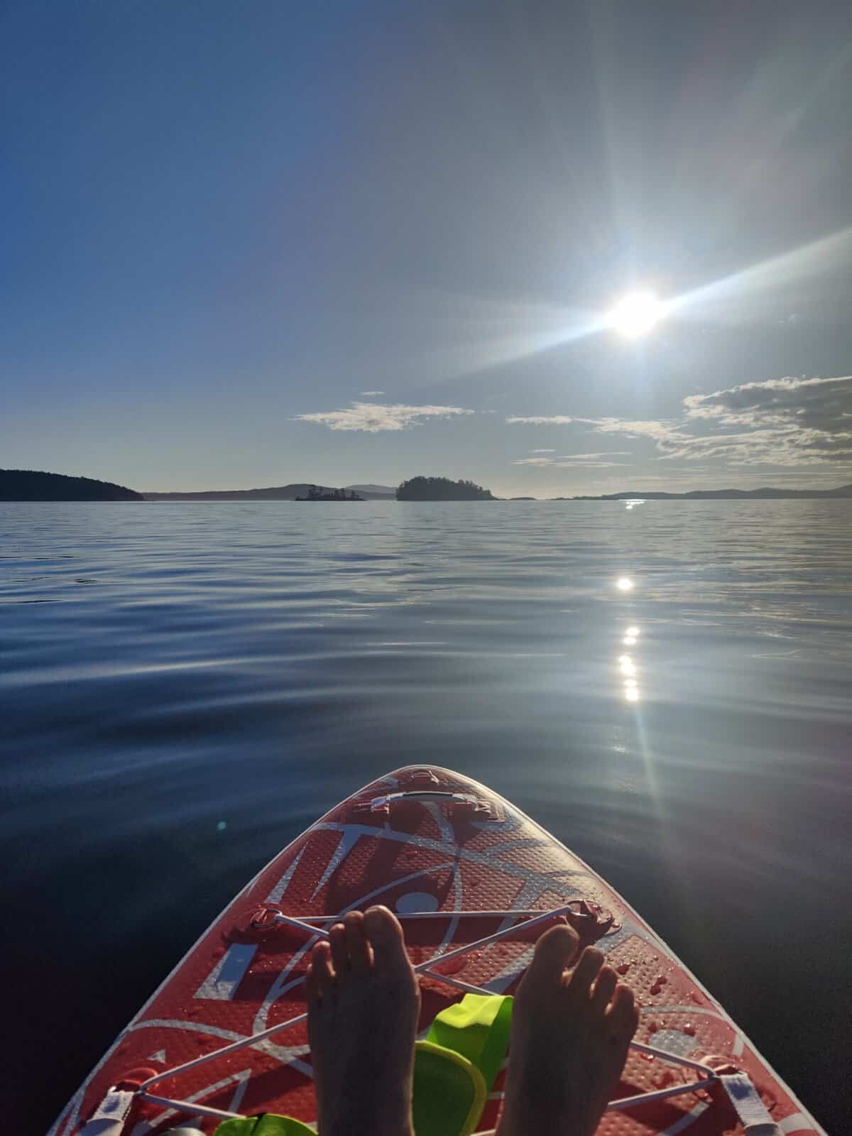 **View from a paddleboard on calm water at sunset, with distant islands and sunlight reflecting across the surface.**
