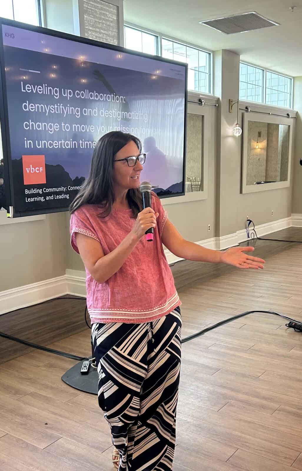 A woman holding a microphone speaks to an audience in a bright, modern room. Behind her, a large presentation screen displays a talk about collaboration and navigating change in uncertain times, with the VBCF logo visible.
