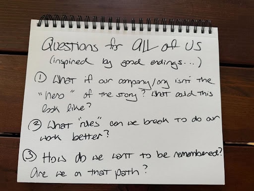 **Alt text:**
A spiral-bound notebook on a wooden table with handwritten text that reads, “Questions for all of us (inspired by good endings…)” followed by three prompts:
1. “What if our company/org isn’t the ‘hero’ of the story? What does this look like?”
2. “What ‘rules’ can we break to do our work better?”
3. “How do we want to be remembered? Are we on that path?”