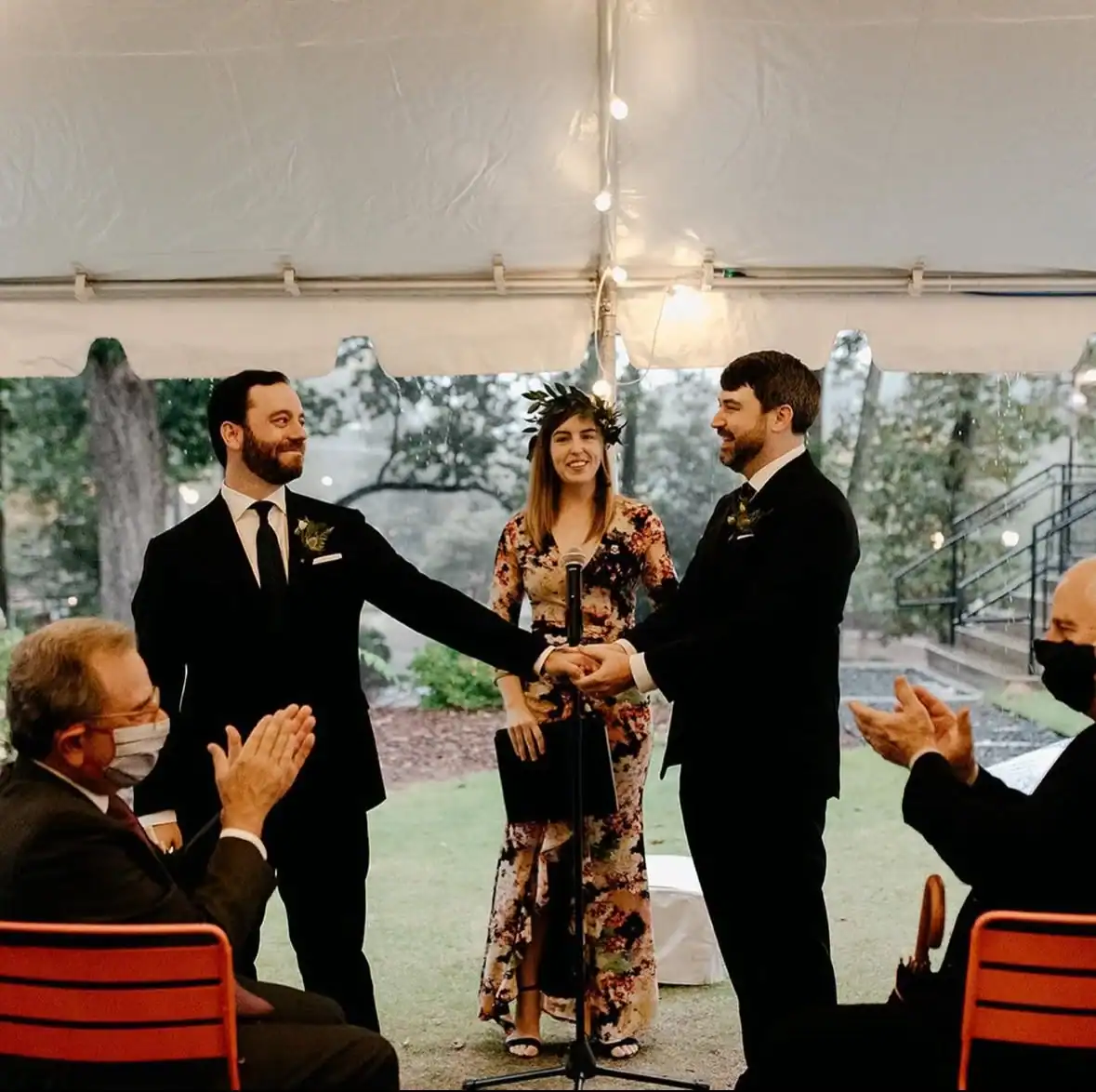 **Alt text:**
Two grooms hold hands during a wedding ceremony under a tent, with an officiant speaking and guests clapping nearby.
