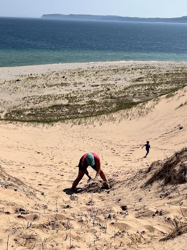 A person crouches on a sandy dune above a beach, with a calm blue ocean and shoreline in the background.
17:03