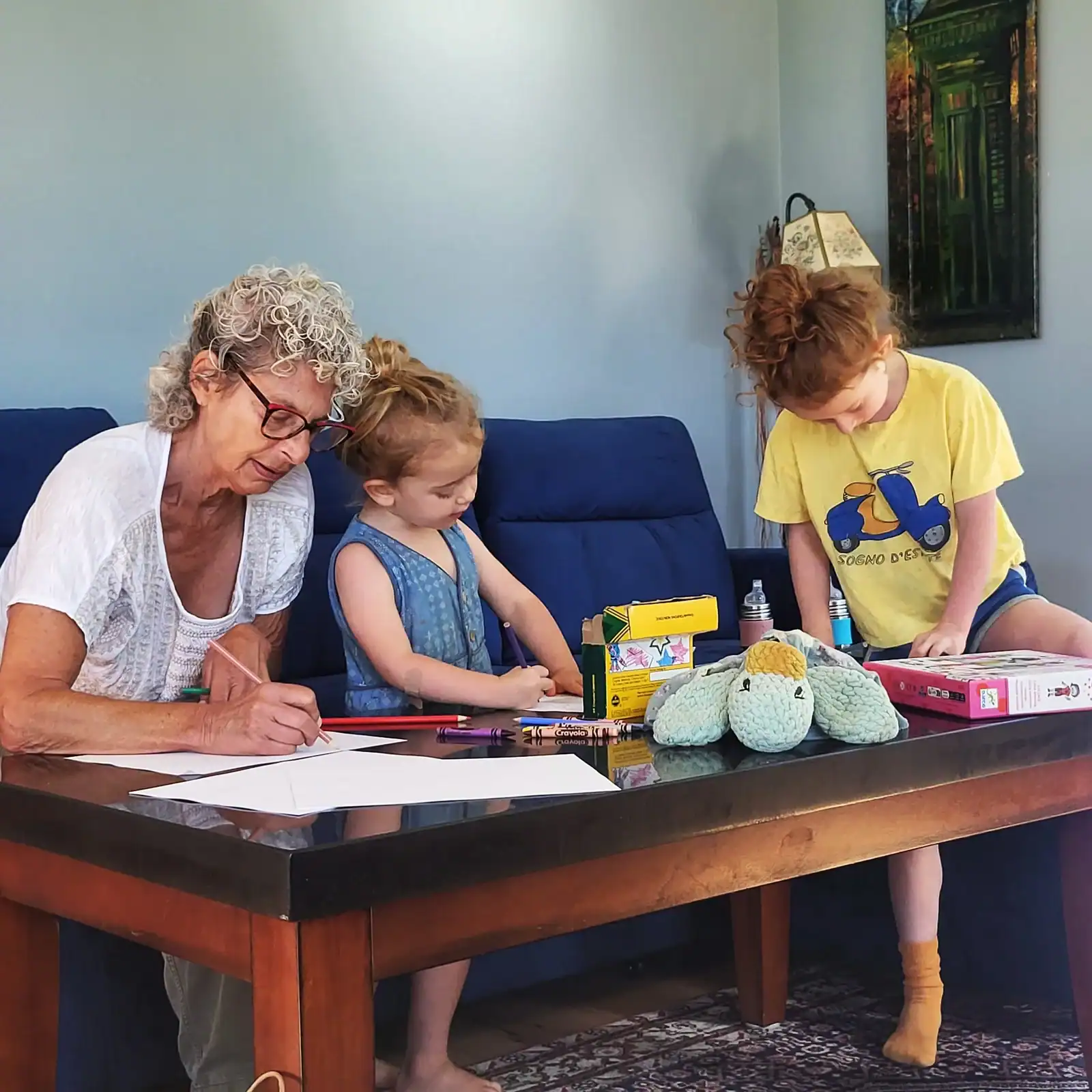 An older woman with curly gray hair and glasses sits at a coffee table helping two young children draw. The children focus on coloring with crayons while a stuffed toy and art supplies are spread across the table, creating a warm, creative family moment.
