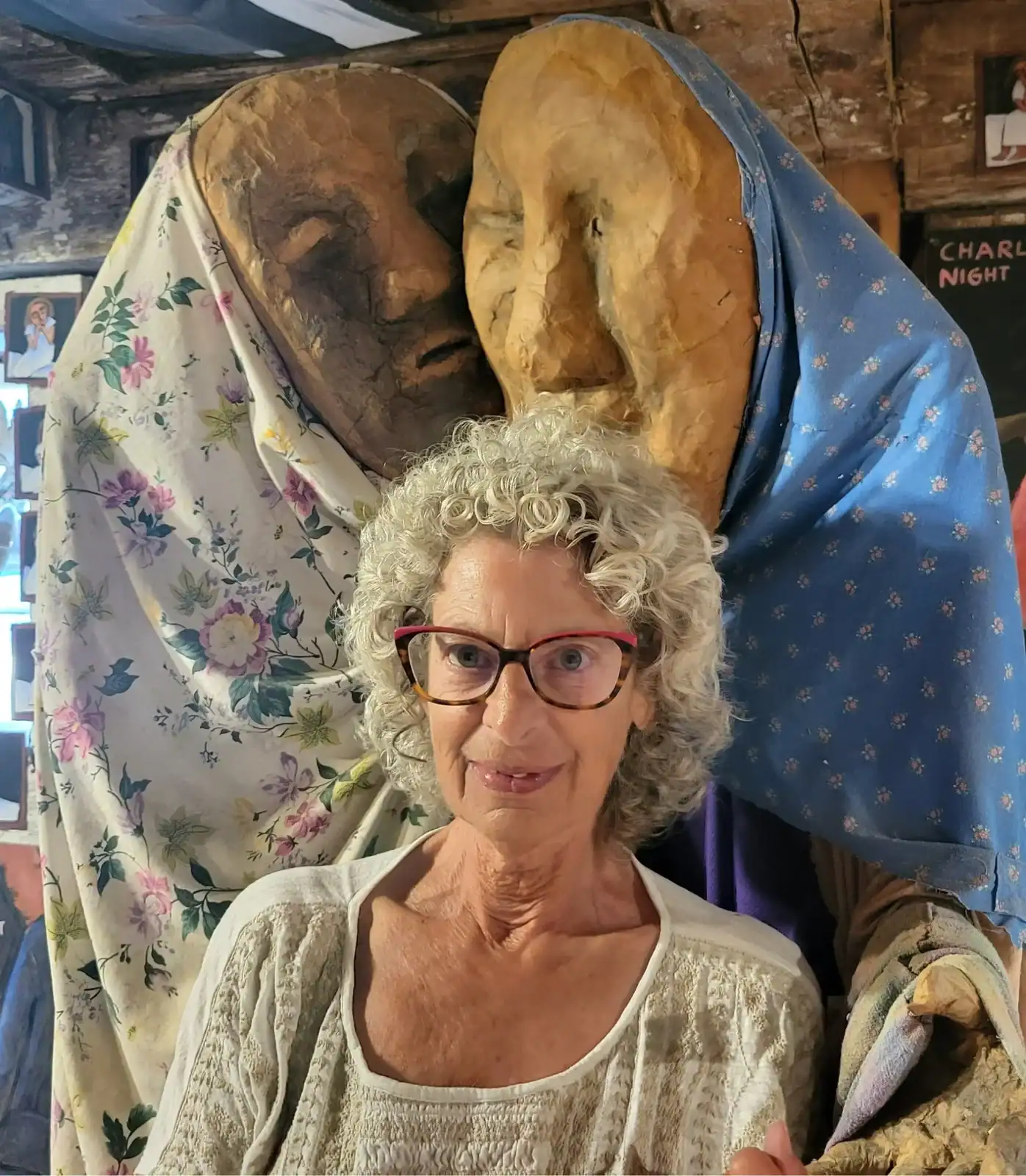 **Portrait of an older woman with curly gray hair and glasses, standing in front of large handmade sculptural figures draped in patterned fabric in a rustic studio setting.**
