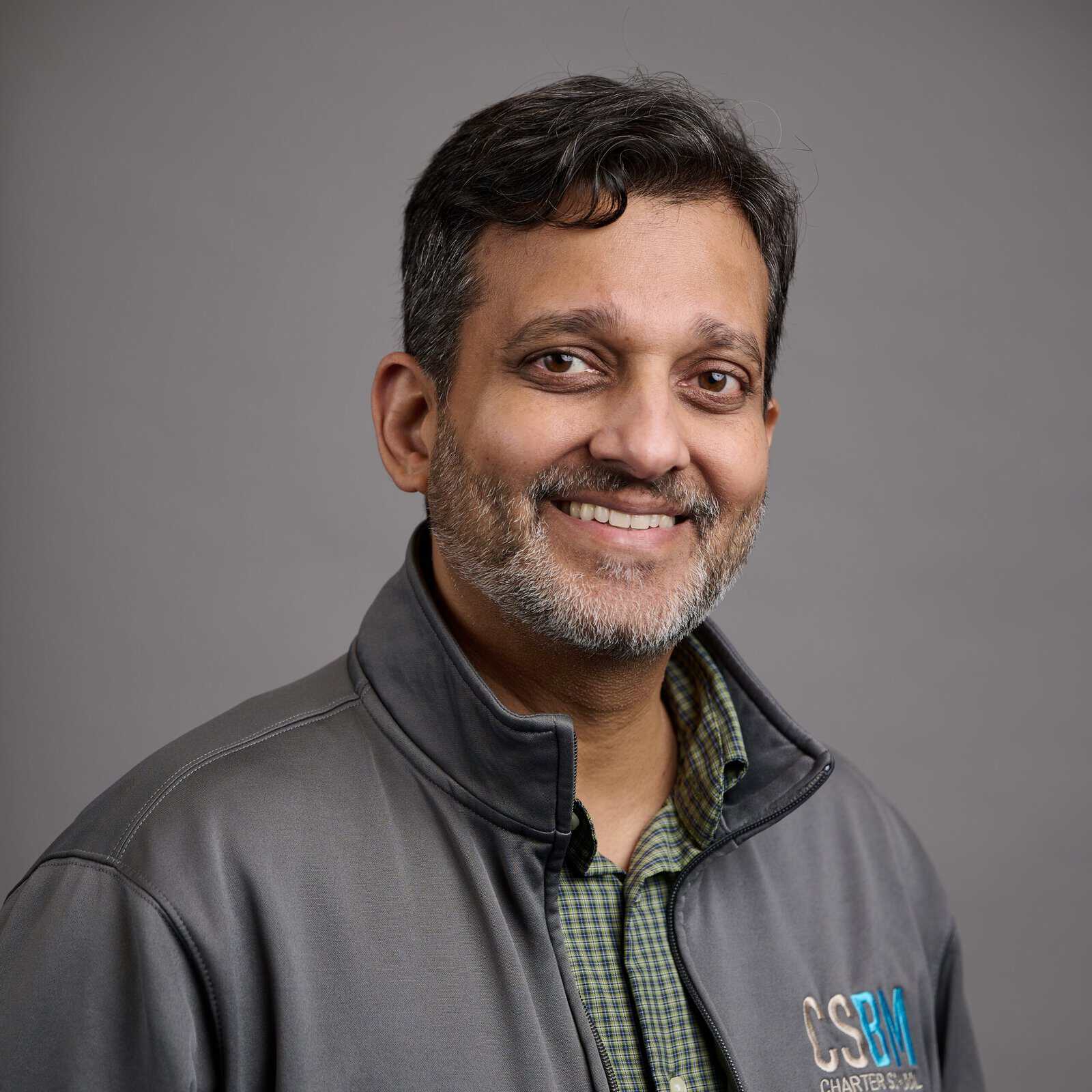 Professional headshot of a smiling man with short dark hair and light stubble, wearing a gray zip-up jacket over a green checkered shirt, against a neutral gray background.