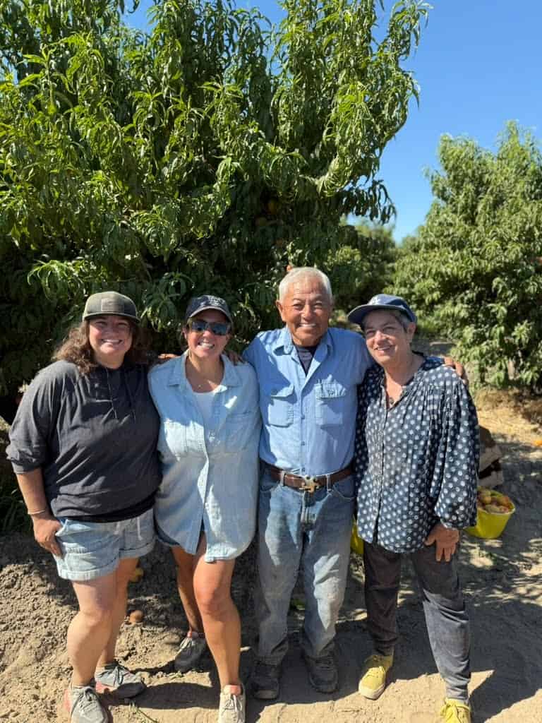 Here's alt text for the image: **Four people smiling together in a peach orchard on a sunny day, with fruit trees and a yellow harvest bucket in the background.**