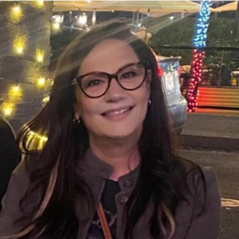 **“Smiling woman wearing glasses, sitting outdoors at night with warm string lights and festive decorations in the background.”**