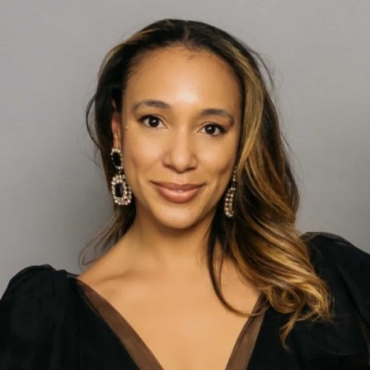 Professional headshot of a woman with long highlighted hair, wearing a black top and statement earrings, smiling gently against a neutral gray background.