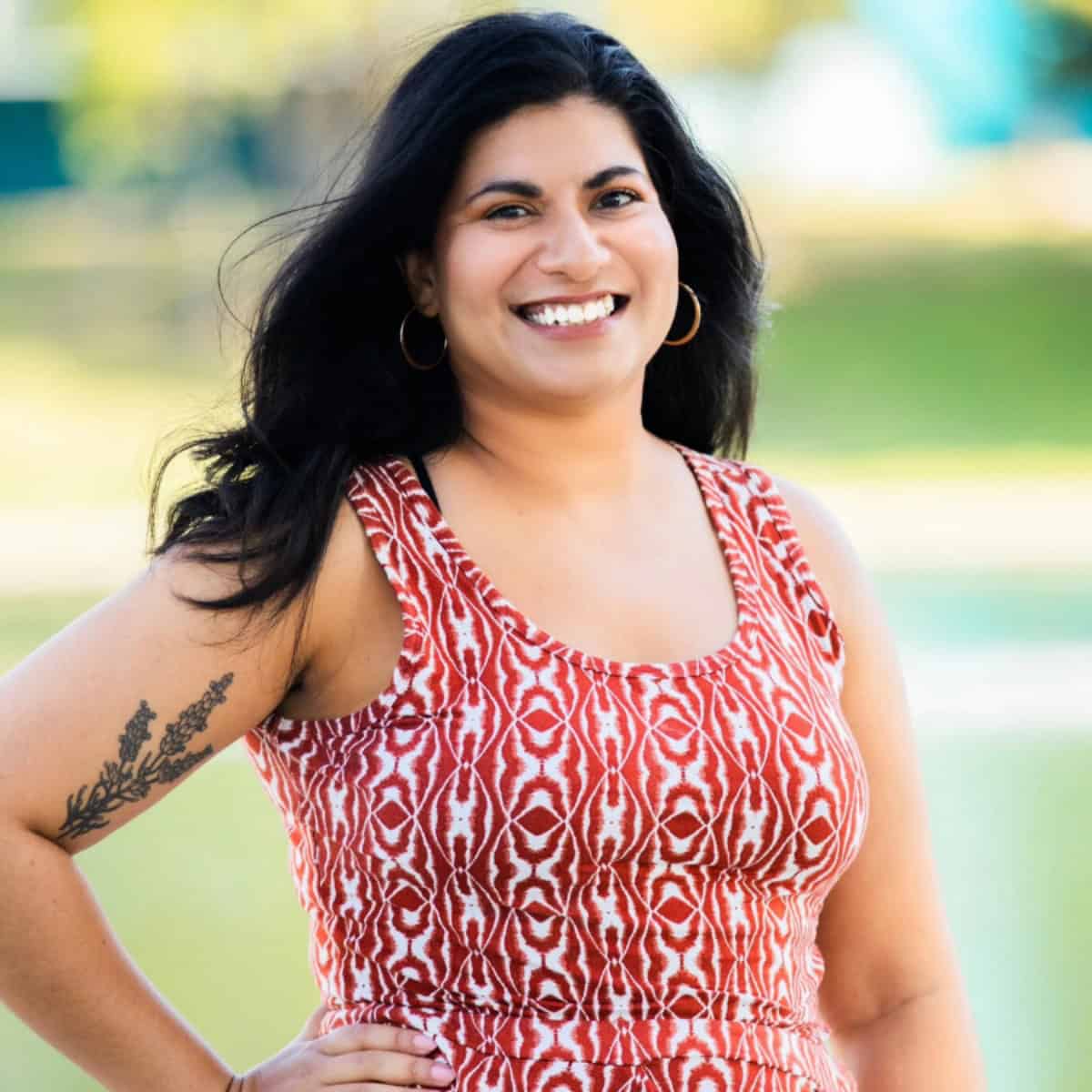 A woman with long, dark wavy hair and gold hoop earrings smiling outdoors. She is wearing a sleeveless red and white patterned dress and has a floral tattoo on her right upper arm. The background is a soft-focus green landscape.
