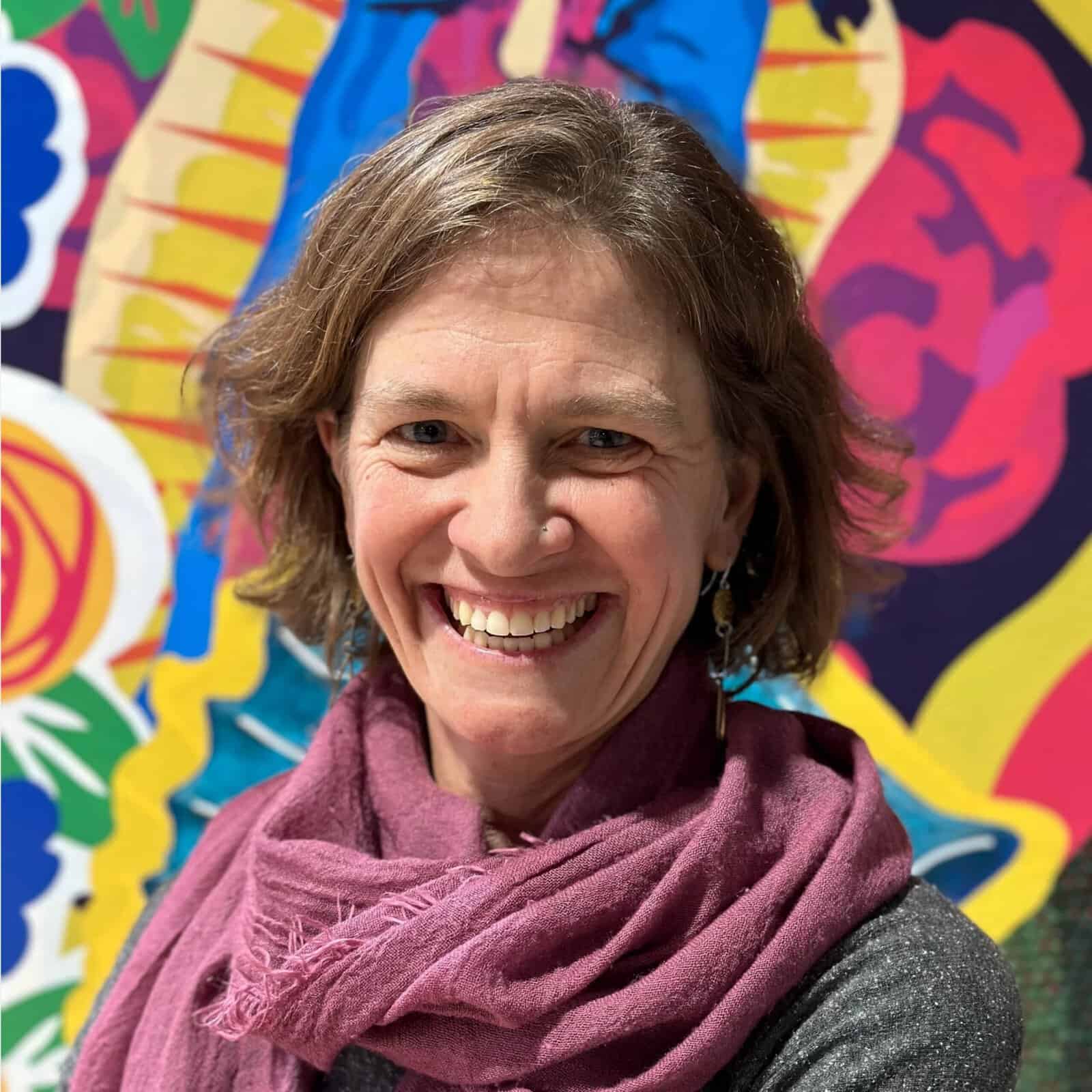 Smiling woman wearing a burgundy scarf, standing in front of a colorful artistic mural.