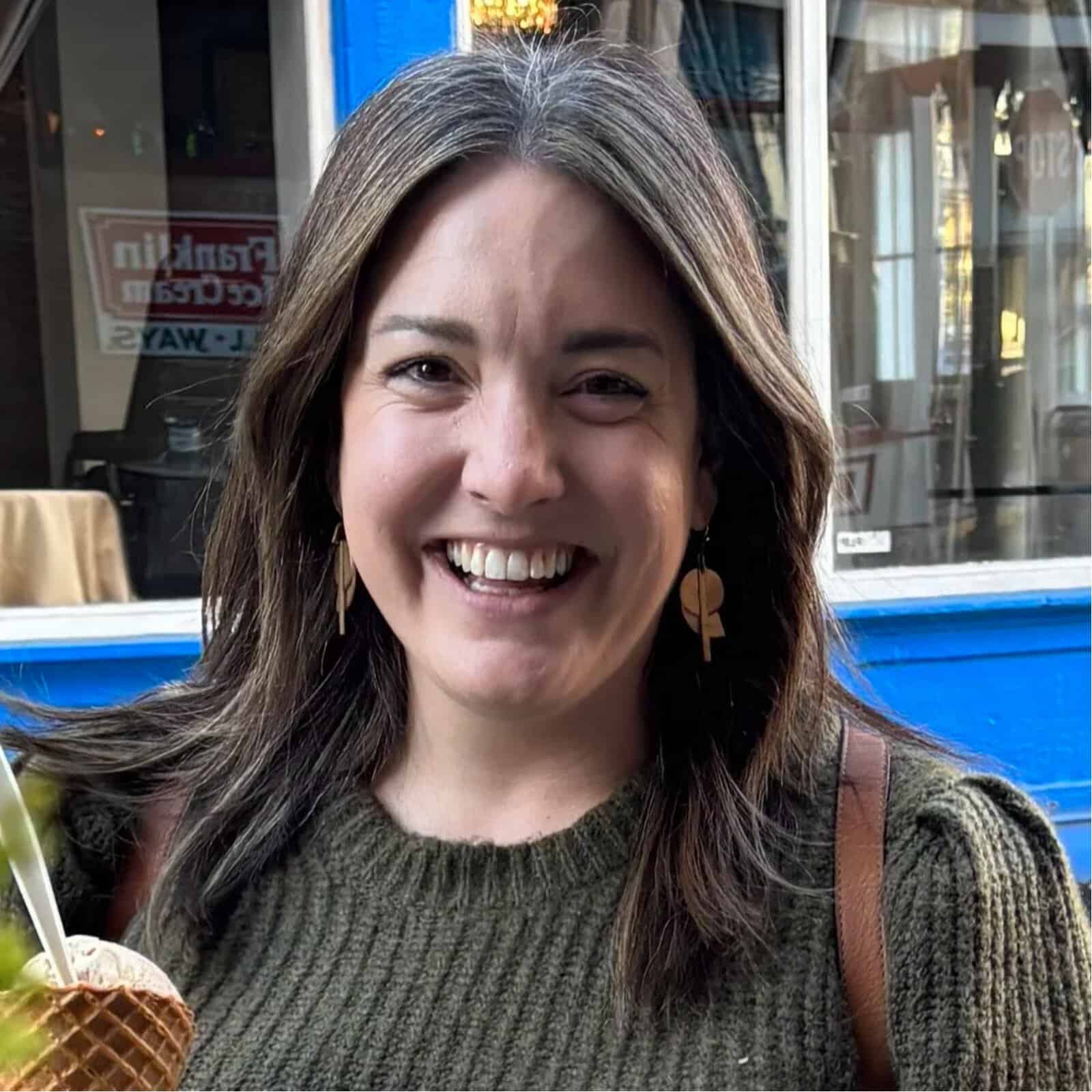 Mujer sonriente al aire libre, con suéter verde y aretes dorados, sosteniendo un helado frente a una fachada azul.