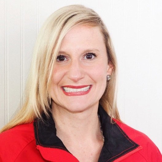 Professional headshot of a smiling woman with long blonde hair wearing a red jacket with a black collar, posed against a light neutral background.