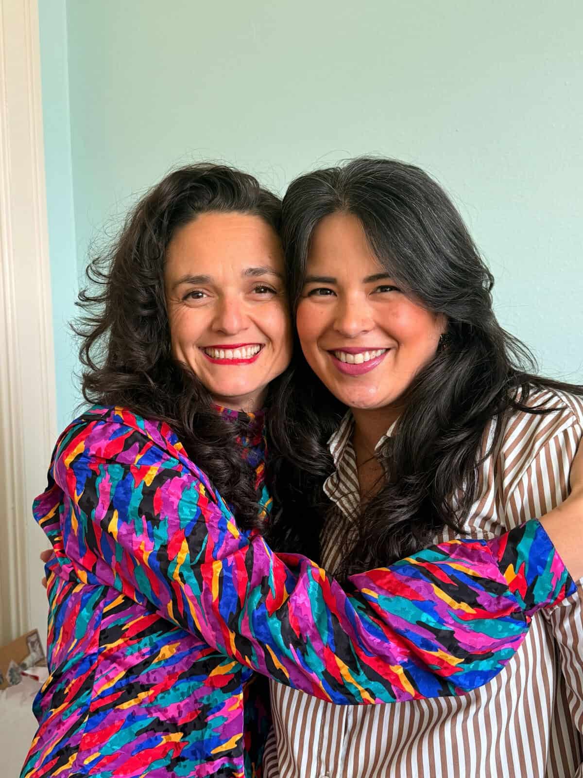 Two women smiling and hugging, standing close together against a soft green wall, conveying warmth and connection.