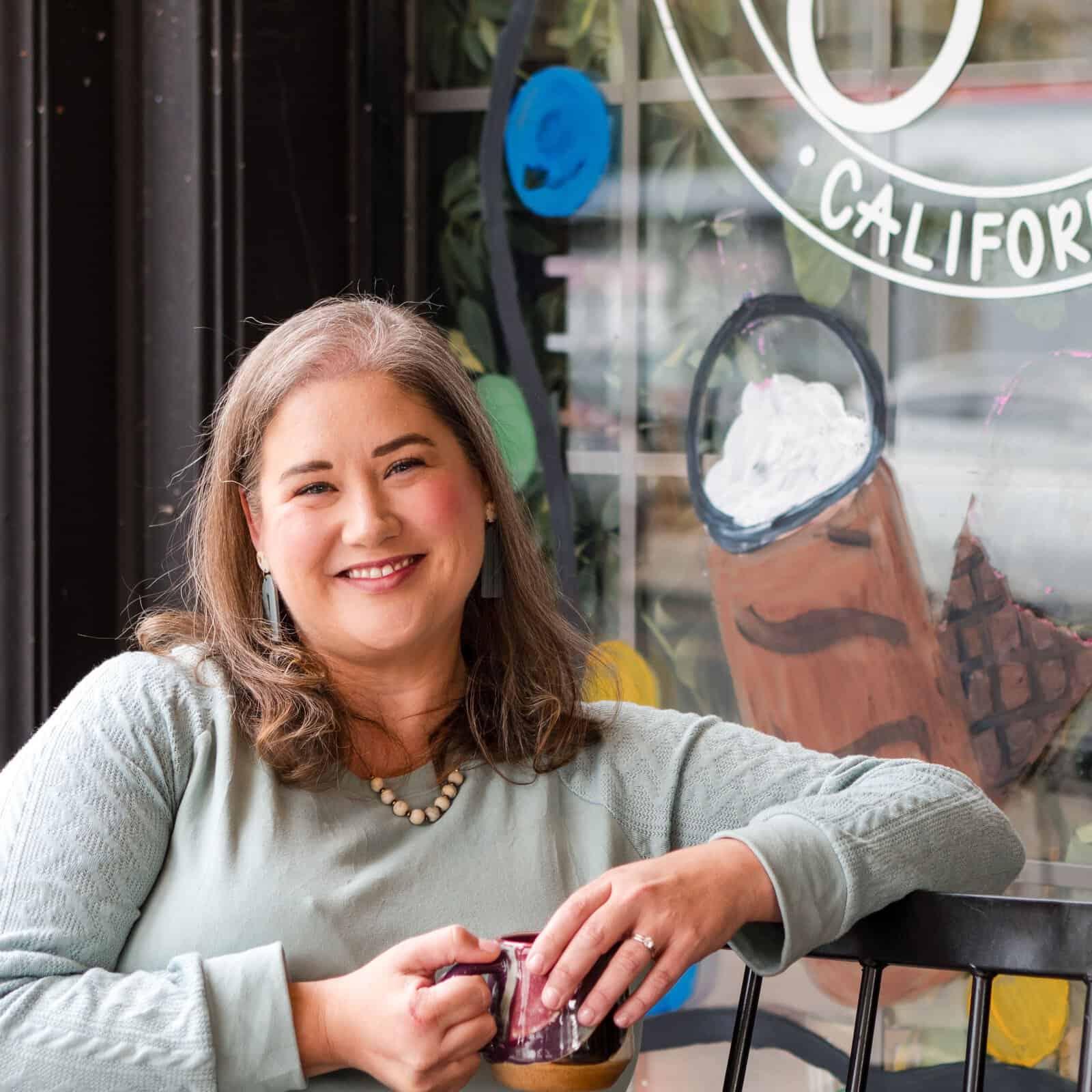 Smiling woman sitting at a café table, holding a mug, with a colorful storefront window and artwork in the background.